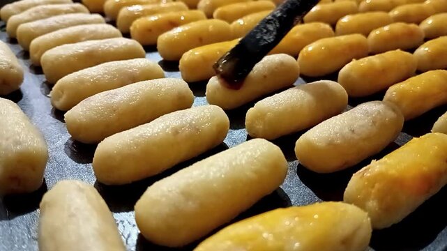 Close view of rows of pale oblong dough pieces arranged on a dark baking tray, with a brush applying glossy liquid to several pieces, showing fine surface texture and even spacing.