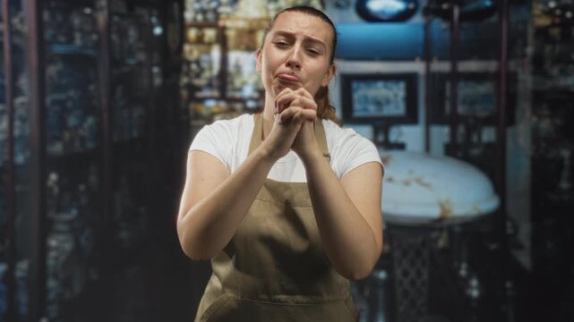 Woman clasping hands at a cafe counter, apron and white tshirt visible, scrunchie in hair, metal roaster and shelves behind; anxiety pleading.