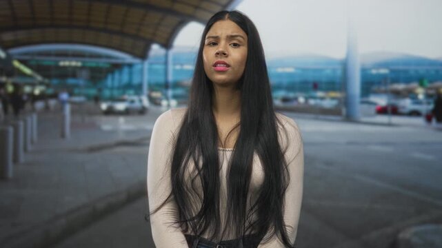 Woman arms crossed with fingers pointing in opposite directions at airport entrance with cars behind; uncertainty.