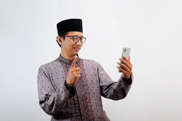 An Asian Muslim man smiles happily while sending a message using his mobile phone, isolated on white background