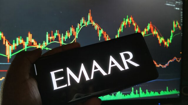Hand holding smartphone with Emaar logo in front of stock market graph display