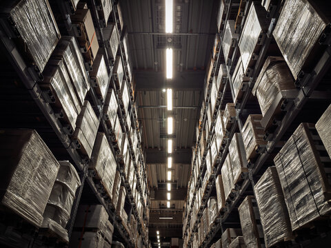 Low angle view of tall shelves stacked with wrapped goods and boxes in a modern logistics warehouse, a symbol of exports, imports, and inventory supply management.
