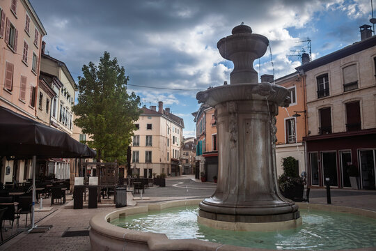 Fountain in Place du 23 Aout 1944 square in Bourgoin-Jallieu, France, with pedestrian plaza, cafe terraces and pastel buildings under cloudy sky in a Dauphine town center, a typical french city.