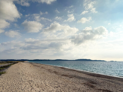 Wide sandy shoreline at Plage de l'Almanarre on Route du Sel in Hyeres, France, with calm Mediterranean water, distant hills of presqu'ile de giens and clouds creating a coastal sky and open seascape.