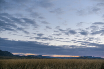 Fototapeta premium Epic cosmic sunset with glowing clouds over a dry yellow steppe and fields in East Kazakhstan. Idyllic rural landscape with distant mountain peaks on the horizon.