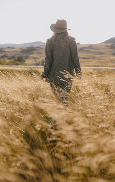 Blonde woman in hat and stylish blazer standing in golden autumn field at sunset. Back view of girl in Kazakhstan steppe with mountains. American west aesthetic, wild nature and freedom concept.