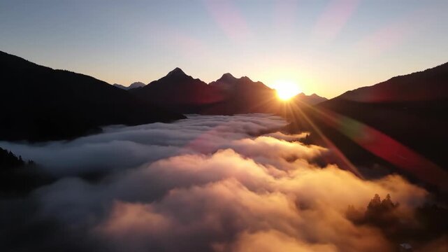 A sunrise with sunbeams peers over mountain peaks and blankets valley in swirling warmcolored cloud cover