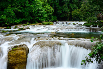 Cascading tiered waterfalls flow over layered limestone steps, forming a series of crystal clear, emerald pools within the lush Xiaoqikong scenic area.