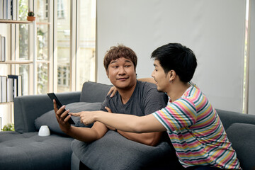 Obraz premium Two Asian LGBTQ men sitting close on sofa, smiling and pointing at a tablet while making eye contact. The moment reflects fun interaction, friendship, and digital lifestyle at home