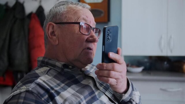 Close-up of elderly man in glasses using his smartphone to record voice message while sitting in cozy home kitchen. Modern technology, communication, and elderly people adapting to digital life.