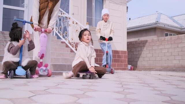 Young sisters ride scooters in paved yard of house while their mother supervises. Concept of childhood, active leisure, safety, family bonding and outdoor play. Girls riding scooters in yard under m