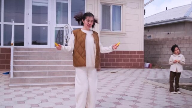 Asian mother jumping rope in front of house while children watch and play around. Authentic everyday lifestyle moment, natural behavior captured indoors, cinematic shot emphasizing real life emotion