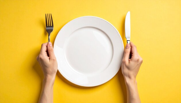 Human hands grasp fork and knife, poised above a clean, empty white plate against a vibrant yellow backdrop, conveying readiness for food, diet concepts, or dining experiences