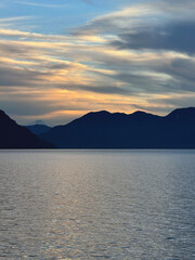 Peaceful sunset over a calm lake with dark mountain silhouettes on the horizon. Soft golden and blue clouds in the sky, minimalistic natural landscape with copy space and no people.