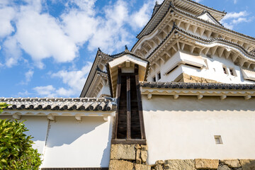 Upward perspective of ornate white turret and layered tiled walls at Himeji Castle, featuring intricate wooden details and stone foundation beneath a bright sky with scattered clouds. © Florent
