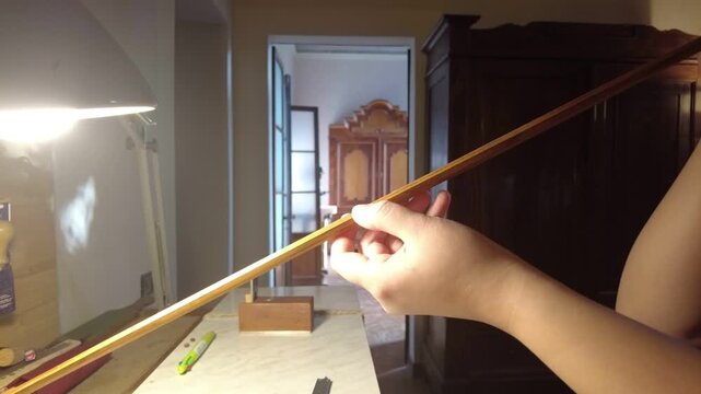 Close-up of a woman artisan inspecting alignment and camber of a pernambuco violin bow stick by looking along shaft on a luthier bench, illustrating precision control in string instrument making