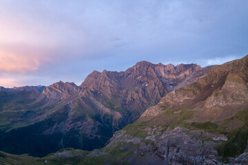 Evening light casts a soft purple hue across the rugged peaks and layered rock faces of the Pyrenees, with gentle clouds drifting above the wild alpine landscape.