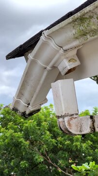 Medium shot view of a wet white rain gutter with residual water droplets hanging off the edge, attached to a dark grey tiled roof on a residential house. Overcast post-rain atmosphere. 4K ProRes 30fps