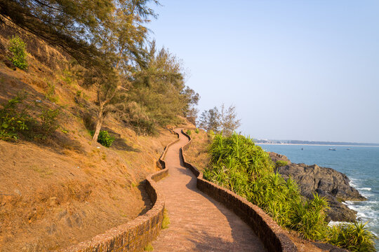 Curved stone walkway hugs the hillside above rocky cliffs and overlooks the Arabian Sea at Bekal Fort, India. Warm sunlight casts gentle shadows on the textured path and surrounding greenery.