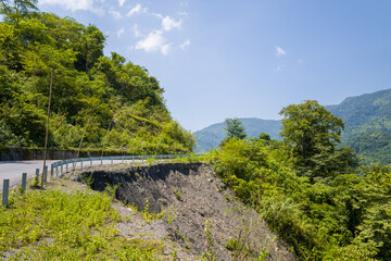 Obraz premium A steep landslide exposes dark soil and rocky earth beside a curving asphalt road in the green hills near Lai Chau, Vietnam. Bright sunlight enhances the vivid foliage and dramatic slope under a clear