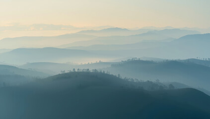 Obraz premium Misty layered mountain ridge at serene sunrise with soft pastel light and distant trees
