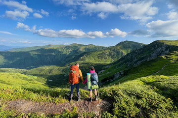 Back view of two hikers, holding hands, stand on trail overlooking mountain landscape. Tourists equipped with backpacks and trekking poles. Rolling hills, rocky outcrops, bright blue sky with clouds. © anatoliy_gleb
