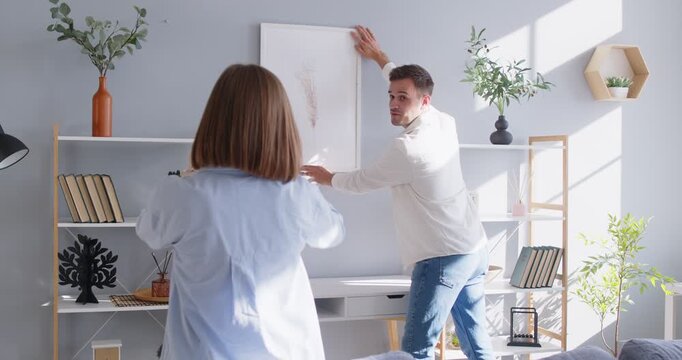 Couple hanging art picture on home wall to organize cozy living room interior together. Man holding frame to hang decor with help of woman, people planning domestic design together with discussion
