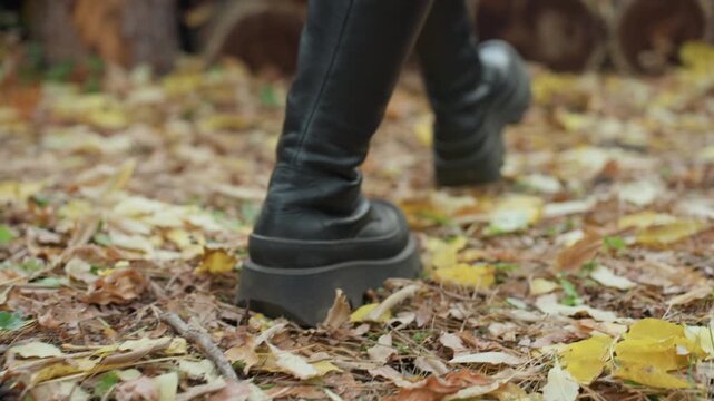 leather boots walking on leaf covered forest floor, closeup of woman in black leather boots stepping through crunchy autumn leaves beside stacked logs, moody overcast lighting, textured bark