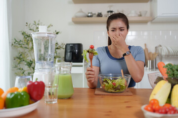 Woman grimacing holding her nose disliking healthy salad in kitchen