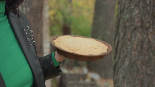 Woman holding homemade pie outdoors wearing leather jacket and green turtleneck, rustic tin plate with golden crust, brick cottage and pine trees in background, closeup on hand and pie, cozy moody