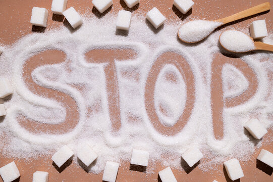Letters STOP written on scattered refined sugar and square sugar cubes on brown background. Concept of high sugar consumption, health problems, excess weight, diabetes. Sucrose, simple carbohydrates