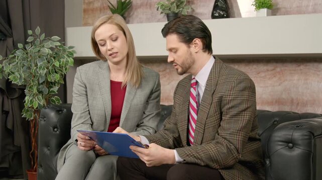 Confident attractive middle aged businesswoman in elegant formal wear molesting shocked handsome male business colleague, making inappropriate proposal and touching hand at office.