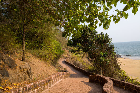 Winding stone walkway bordered by lush greenery leads down toward a sandy beach and calm sea in Bekal, India. Warm sunlight filters through leafy branches, casting soft shadows on the textured path.