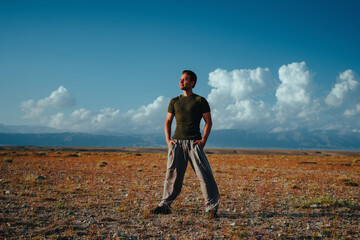 Young european man standing in steppe with hands in pockets, looking at the sun happily