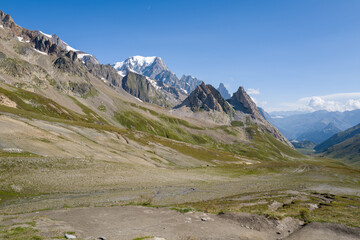 Obraz premium Expansive mountain valley at Col de la Seigne featuring rugged rocky ridges, green slopes, and the snow-capped summit of Mont Blanc beneath a clear blue sky. The landscape is open and sunlit