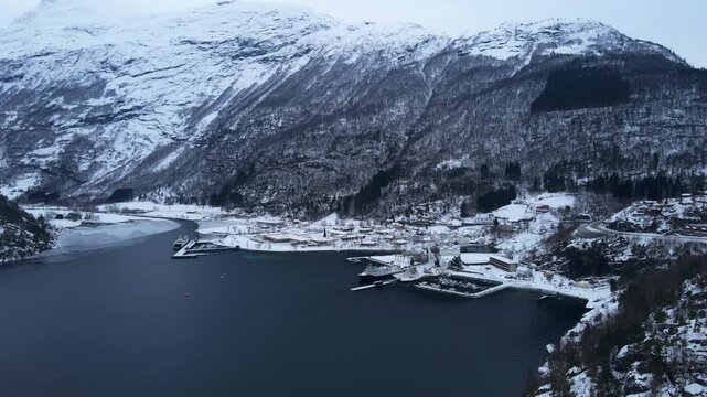 Drone shot of Hellesylt, a small town in Norway surrounded by fjords and tall mountains with snow covering the landscape on a bright winter day