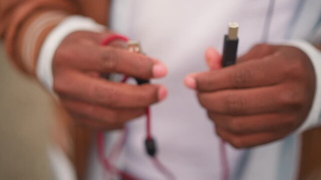 black busker hands plugging red cable for street performance, intimate closeup of USB and jack on braided cord, casual jacket and denim, testing gear and tuning before busking set