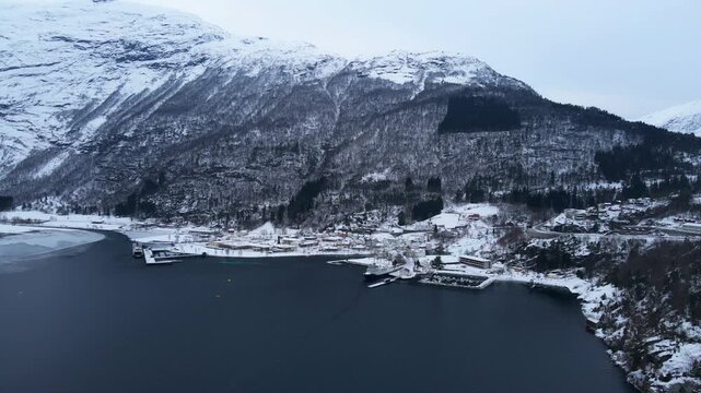 Drone shot of Hellesylt, a small town in Norway surrounded by tall mountains and fjords with snow covering the landscape on a bright winter day in Norway