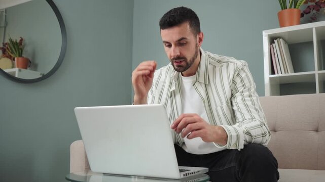 A concerned man seated on a comfortable couch is dealing with stress and frustration while using a laptop in a stylish, contemporary living space, showcasing a relatable moment of modern life