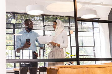 Diverse coworkers wearing hijab reviewing files, holding phone in office behind glass partition © wavebreak3
