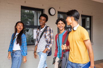 Diverse student friends walking and talking along campus corridor with backpacks and tablet