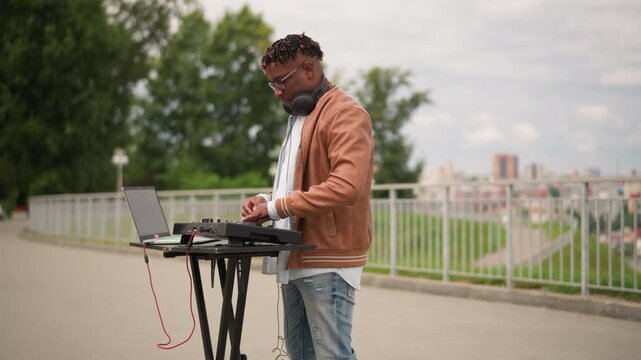 black DJ performing outdoor keyboard set in park with laptop and midi controller on stand, brown jacket, headphones, city skyline background, railing, daytime vibe, upbeat mixing, busker energy,