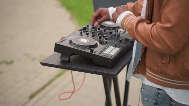 dj controller on outdoor stand mixing, brown jacketed musician adjusts knobs on compact mixer beside laptop, red cable dangling, pavement backdrop, intimate street performance vibe, focused hands