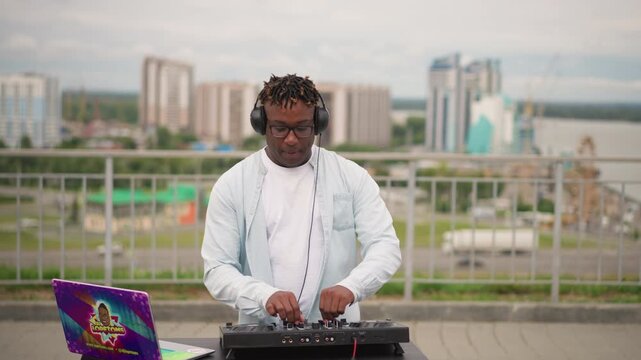 black deejay rooftop skyline mixing mellow sunset set with focused producer energy solo artist concentrates over controller and headphones, close up on hands adjusting knobs, sampling loops