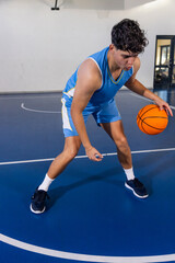 Fototapeta premium Teenage male athlete dribbling orange basketball on court in gym near hoop, glass-panel door