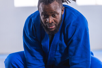 African american man kneeling on blue padded mat inside martial arts studio in blue gi