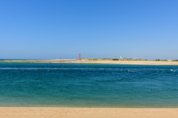 Fototapeta premium Vivid blue tidal water flows along a wide sandy shoreline under a clear sky at Barre d'Etel in Brittany. Distant navigation markers and a white signal tower add structure to the bright, tranquil