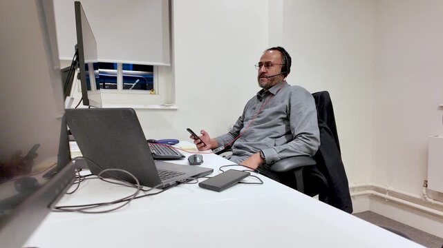 South Asian Man Talking On Headset Swiveling In Chair Holding Phone At Desk With Laptop And Dual Monitors In Office Setting Call Center Concept.