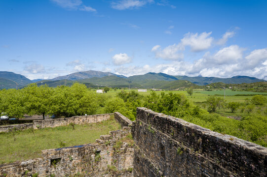 Weathered stone ramparts rise above lush fields and trees, with rolling mountains and a bright blue sky in the background near Ainsa, Spain. The scene combines historic textures with vibrant rural