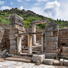 Fototapeta premium Weathered stone ruins and steps of ancient Ephesus, a historic site in Selcuk, Turkey, set against green hills and a cloudy sky